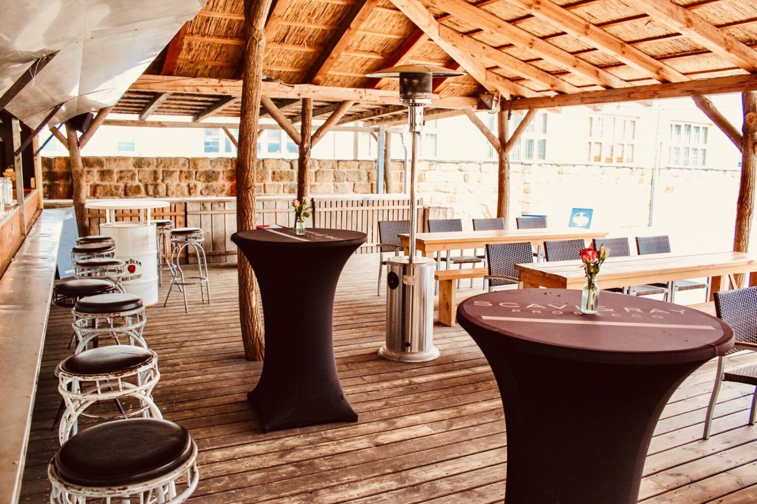 Ein helles, querformatiges Foto zeigt eine Bar im Freien auf einer Holzterrasse. Im Vordergrund stehen zwei Stehtische mit schwarzen Hussen. Auf den Tischen stehen kleine Vasen mit roten Rosen. Links im Bild stehen mehrere Barhocker mit braunen Ledersitzflächen. Im Hintergrund sind Holztische und Rattanstühle zu sehen. Die Terrasse hat ein Dach aus hellen Holzlatten und dunkle Holzbalken. Auf der Terrasse steht auch ein silberner Heizstrahler. Im Hintergrund ist ein Gebäude mit Fenstern zu erkennen. Das Foto vermittelt eine einladende, entspannte Atmosphäre für eine Bar im Freien mit Sitzgelegenheiten. Firmenfeier am Citybeach