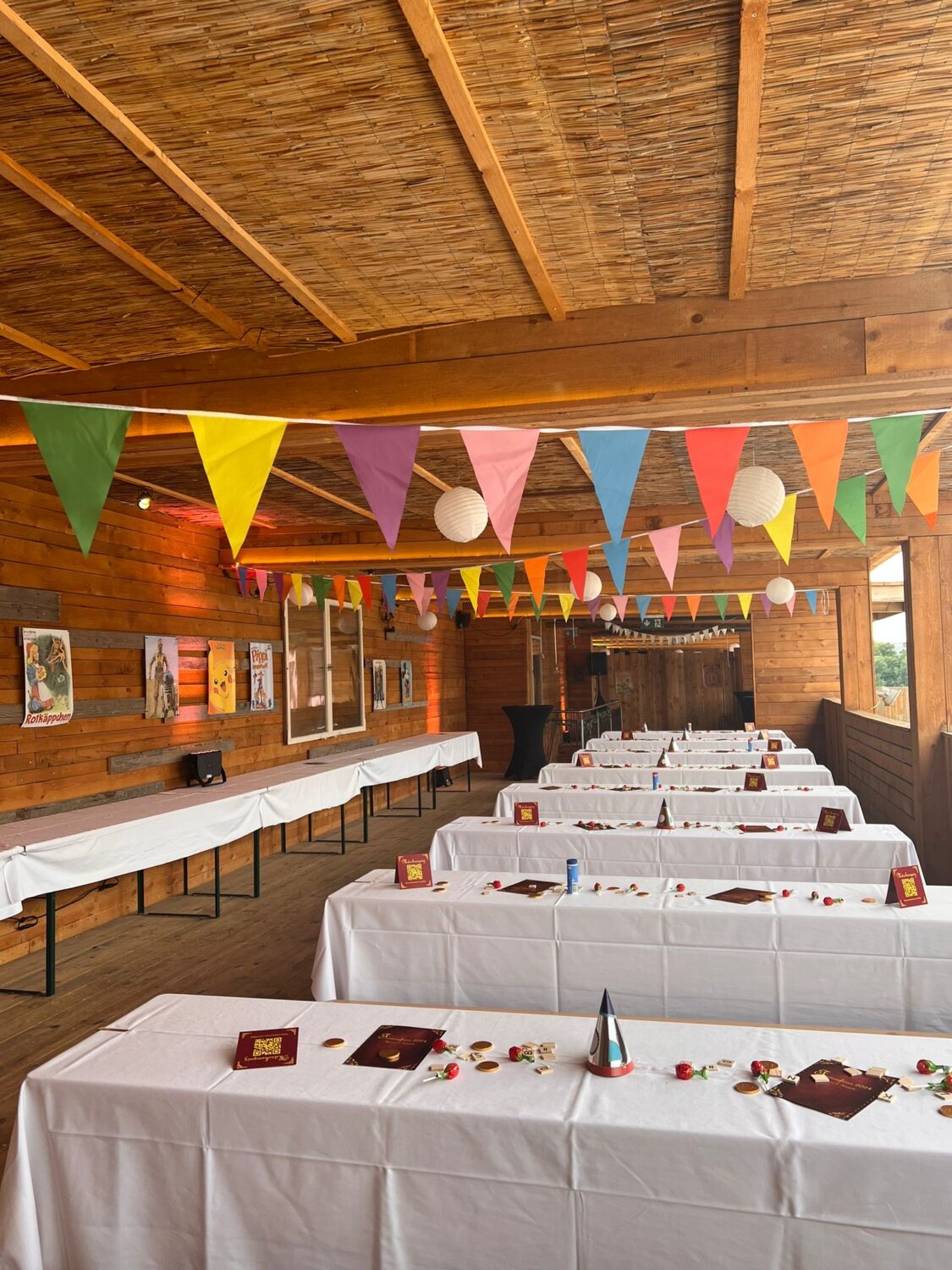 Ein helles, hochformatiges Foto zeigt eine Art Loggia mit festlich gedeckten langen Tafeln. Die Loggia hat ein Dach aus hellen Holzlatten und Wände aus dunklen Holzbrettern. An der Decke hängt eine bunte Wimpelgirlande mit dreieckigen Wimpeln in verschiedenen Farben. Im Hintergrund hängen weiße Lampions. Im Vordergrund stehen mehrere lange Tafeln mit weißen Tischdecken, dekoriert mit roten und goldenen Elementen. Auf den Tischen stehen auch rote Kartenhalter. Im Hintergrund sind Bilderrahmen an der Wand zu sehen. Der Boden besteht aus hellen Holzdielen. Das Foto vermittelt eine festliche, einladende Atmosphäre für eine Veranstaltung im Freien. Firmenfeier am Citybeach