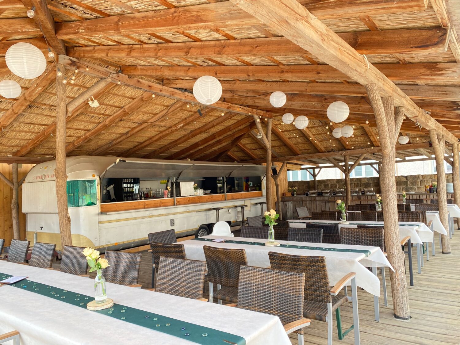 Ein helles, querformatiges Foto zeigt einen großen, offenen Holzpavillon mit festlich gedeckten langen Tafeln einer Firmenfeier am Citybeach. Der Pavillon hat ein helles Holzdach und dunkle Holzbalken. An der Decke hängen mehrere runde, weiße Lampions und Lichterketten. Im Hintergrund steht ein umgebauter, silberner US-Airstreamer, der als Bar dient. Im Vordergrund sind mehrere lange Tafeln mit weißen Tischdecken und grünen Tischläufern zu sehen. Auf den Tischen stehen kleine Vasen mit gelben Rosen. Braune Stühle stehen entlang der Tafeln. Der Boden besteht aus hellen Holzdielen. Das Foto vermittelt eine festliche, einladende Atmosphäre im Freien mit einer mobilen Bar und dekorierten Tischen.