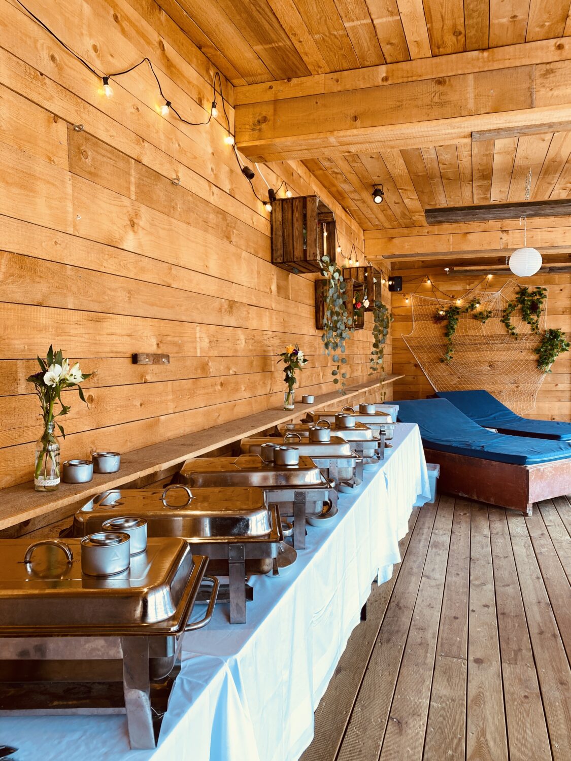 Ein helles, hochformatiges Foto zeigt eine hölzernen überdachte Terrasse mit einem Buffet einer Firmenfeier am Citybeach. Die Terrasse hat Wände und eine Decke aus hellen Holzbrettern. An der Decke hängen Lichterketten und eine Lampe. An den Wänden hängen hölzerne Kisten, grüne Pflanzen und ein Netz mit Dekoration. Im Vordergrund stehen mehrere Buffet-Tische mit warmen Speisen in silbernen Wärmebehältern. Die Tische sind mit weißen Tischdecken bedeckt. Auf den Tischen stehen auch kleine Vasen mit weißen Blumen. Rechts im Bild stehen hölzernen XL-Liegen mit blauen Matratze. Der Boden besteht aus hellen Holzdielen. Das Foto vermittelt eine einladende, festliche Atmosphäre in einem dekorierten Pavillon mit einem vorbereiteten Buffet.