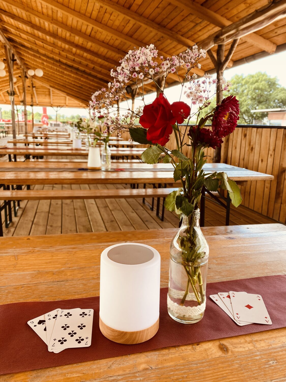 Ein helles, hochformatiges Foto zeigt einen rustikalen Holztisch im Freien auf der Dachterrasse. Auf dem Tisch stehen eine klare Glasvase mit einem Strauß aus roten Blüten und rosa Blütenzweigen, eine weiße zylindrische Lampe mit Holzsockel und zwei Sätze Spielkarten. Der Tisch hat eine dunkelrote Tischdecke. Im Hintergrund sind mehrere lange Holztische und Holzbänke zu sehen. Das Dach der Dachterrasse über den Tischen besteht aus hellen Holzbrettern und dunklen Holzbalken. An der Decke hängen mehrere helle Lampen. Im rechten Bildbereich ist ein Teil eines grünen Baumes zu erkennen. Das Foto vermittelt eine sommerliche, entspannte Atmosphäre im Freien.
