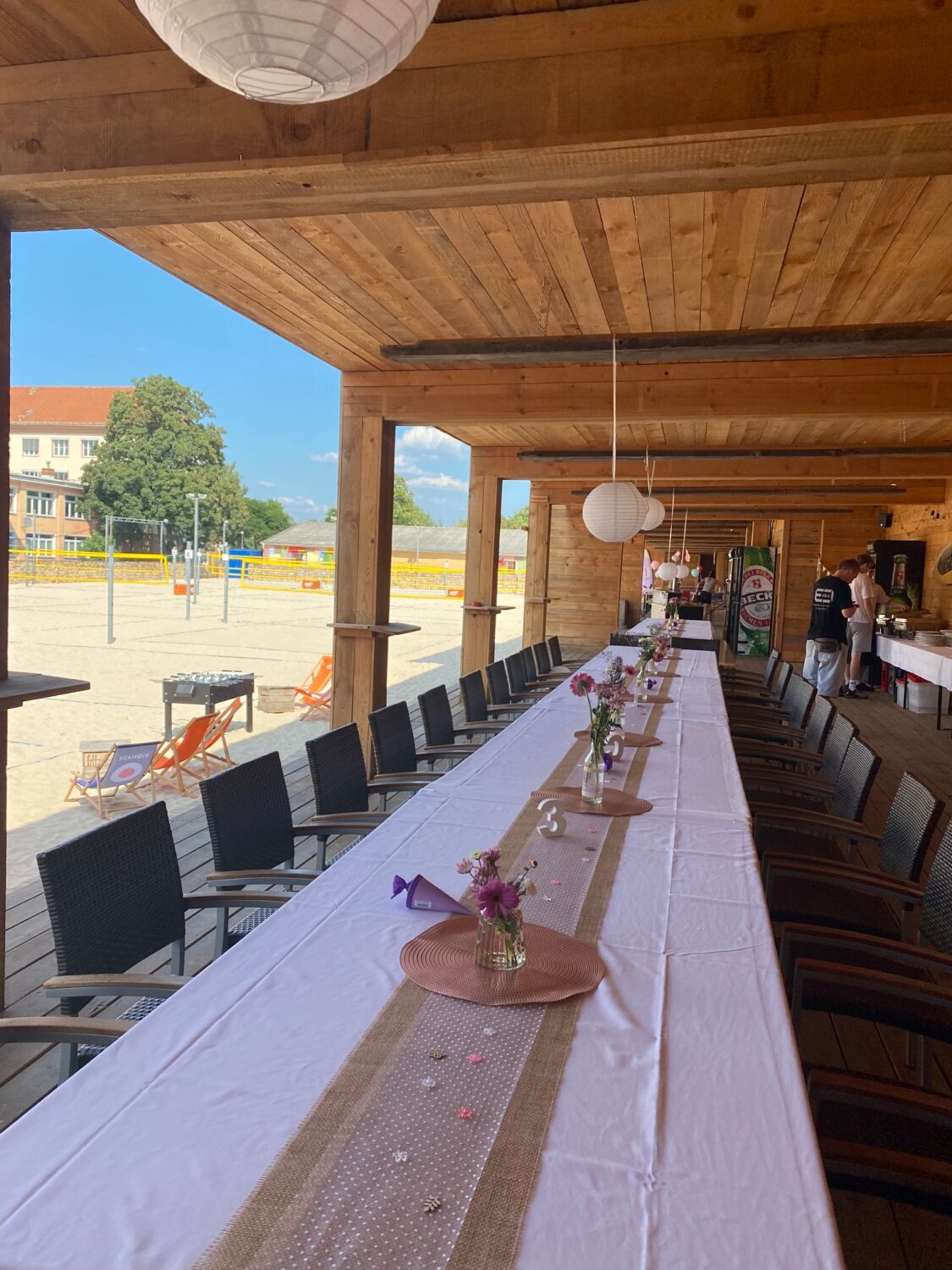 Ein helles, hochformatiges Foto zeigt eine lange, festlich gedeckte Tafel in einer offenen Terrasse einer Firmenfeier am Citybeach. Die Tafel ist mit einer weißen Tischdecke und einem braunen Jute-Tischläufer bedeckt. Auf dem Tisch stehen mehrere kleine Vasen mit lila Blumen und Streudekoration. Im Hintergrund sind schwarze Stühle entlang der Tafel aufgereiht. Die Terrasse hat ein helles Holzdach und dunkle Holzbalken. An der Decke hängen mehrere runde, weiße Lampions. Im linken Bildbereich ist ein Beachvolleyballfeld mit hellem Sand und Netzen zu sehen, sowie Liegestühle und ein Tischkicker. Im rechten Bildbereich stehen zwei Personen an einem Tisch. Das Foto vermittelt eine sommerliche, festliche Atmosphäre im Freien.