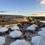 Citybeach aus der Vogelperspektive zeigt den Citybeach Dresden von oben. Zu sehen ist unter anderem die Dachterrasse mit Menschen, die Sandfläche mit Liegestühlen und Sonnenschirmen, sowie die Elbe. Das Bild wurde an einem Sommerabend aufgenommen bei schönstem Sonnenschein.