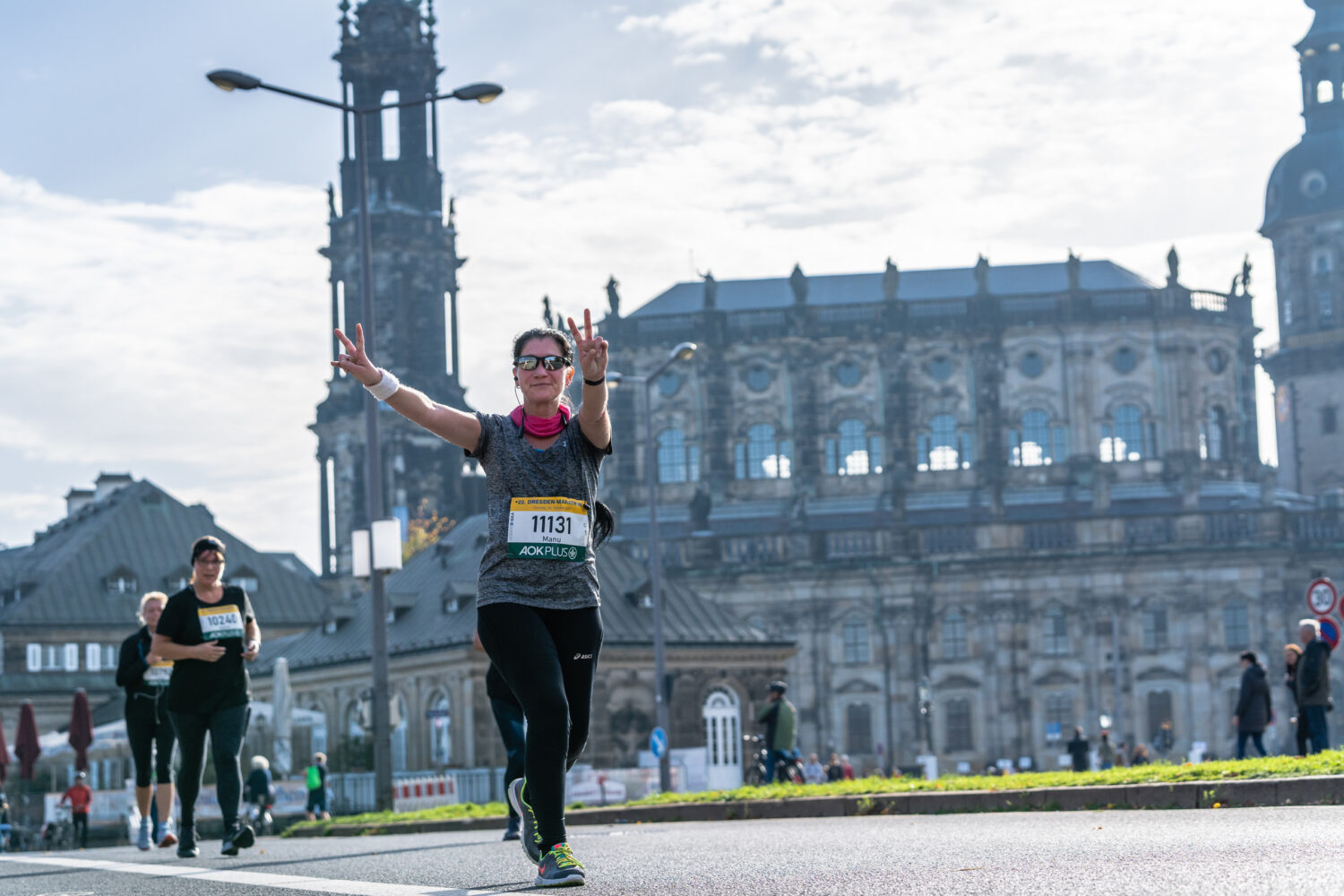 Eine gut gelaunte LĂ€uferin zeigt beim Frauenlauf in Dresden das Victory-Zeichen vor der Kulisse der historischen Hofkirche.