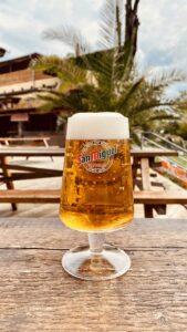 Ein kühles San Miguel Pils in einer Biertulpe mit perfekter Schaumkrone auf einem Holztisch im Citybeach Dresden mit Palme im Hintergrund – Bereich Speisen & Getränke.
